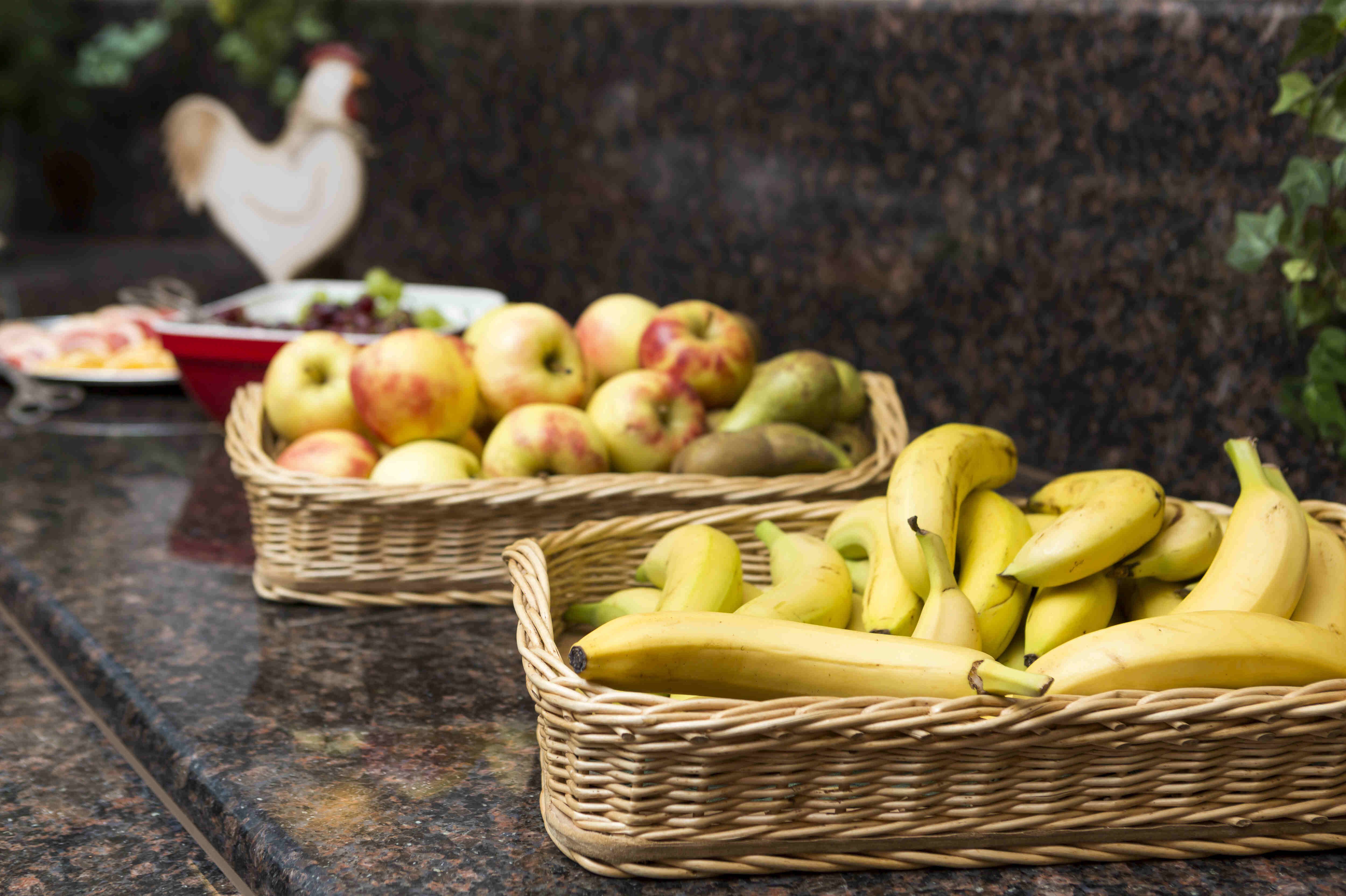 Auf einer glänzenden, dunklen Granit-Arbeitsplatte stehen zwei geflochtene Obstkörbe aus hellem Naturmaterial. Der vordere Korb ist prall gefüllt mit gelben Bananen. Direkt dahinter steht ein zweiter Korb, in dem eine bunte Mischung aus Äpfeln, Birnen und möglicherweise Kiwis liegt. Die Früchte sind sorgfältig arrangiert und wirken frisch und einladend.
