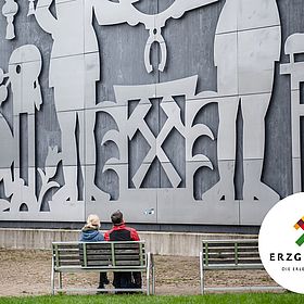 Sportpark Rabenberg EURORANDO 2026 Aussteller: Tourismusverband Erzgebirge; © Tourismusverband Erzgebirge, Dennis Stratmann Paar sitzt auf einer Bank vor einer silbernen Wand mit Metallrelief, das bergmännische Symbole und Figuren zeigt.