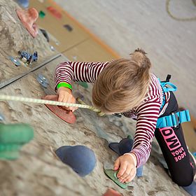 Das Bild zeigt ein Kind beim Klettern an einer Kletterwand. Das Kind ist von oben fotografiert, so dass man es von Kopf bis Fuß sehen kann, während es die Wand erklimmt. Es ist ein junges Mädchen, dessen blondes Haar zu einem kleinen Pferdeschwanz zusammengebunden ist. Sie trägt ein langärmliges rot-weiß gestreiftes Oberteil, schwarze Leggings und einen blauen Klettergurt. Ein Sicherungsseil ist an dem Klettergurt des Kindes befestigt und verläuft nach oben und aus dem Bild heraus.