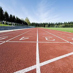 Sportpark Rabenberg: Tartanbahn Das Bild zeigt eine Laufbahn in einem Stadion, die im Freien liegt. Die Laufbahn besteht aus rotem, gummiartigem Material. Es gibt mehrere nummerierte Bahnen, die durch weiße Linien getrennt sind. Die Zahlen sind groß und weiß auf die Bahnen gemalt. Die Perspektive ist so, dass die Bahnen sich in der Ferne zu einem Punkt verjüngen. Links neben der Laufbahn befindet sich eine erhöhte Tribüne aus Beton, die mit einem Metallgeländer versehen ist. Dahinter erstreckt sich eine grüne Rasenfläche mit hohen Bäumen, die einen Wald bilden. Auf der rechten Seite der Laufbahn befindet sich ebenfalls eine grüne Rasenfläche. Im Hintergrund, am Ende der Laufbahn, sind weitere Bäume und ein Gebäude mit kleinen Fenstern zu sehen. Der Himmel ist blau mit ein paar Wolken.