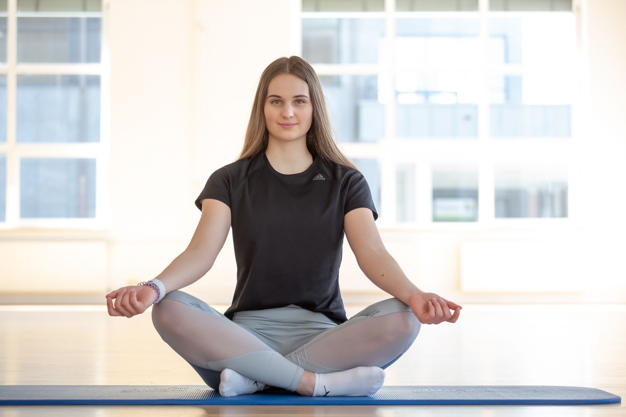 In einem hellen Raum sitzt eine junge Frau im Lotussitz auf einer blauen Yogamatte. Sie trägt ein schwarzes T-Shirt und hellgraue Leggings mit weißen Socken. Sie lächelt leicht, und ihre Arme liegen entspannt auf ihren Knien. Im Hintergrund sind helle Fenster zu sehen.