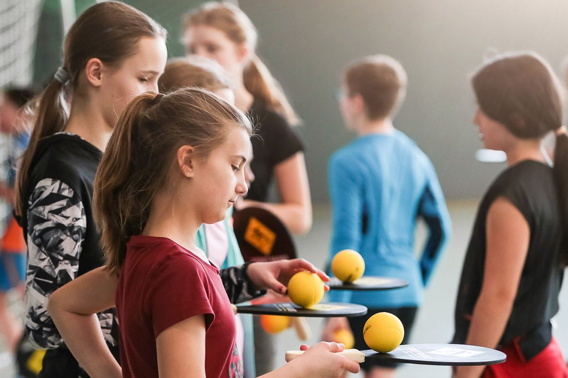 In der Aufnahme ist eine Gruppe von Kindern in einer Sporthalle zu sehen, die ein Ballspiel spielen. Etwa sieben Kinder stehen zusammen. Einige von ihnen halten Schläger und gelbe Bälle. Die Mädchen in den Vordergrund halten ihre Schläger und Bälle und bereiten sich darauf vor, zu spielen. Die Halle ist gut beleuchtet.