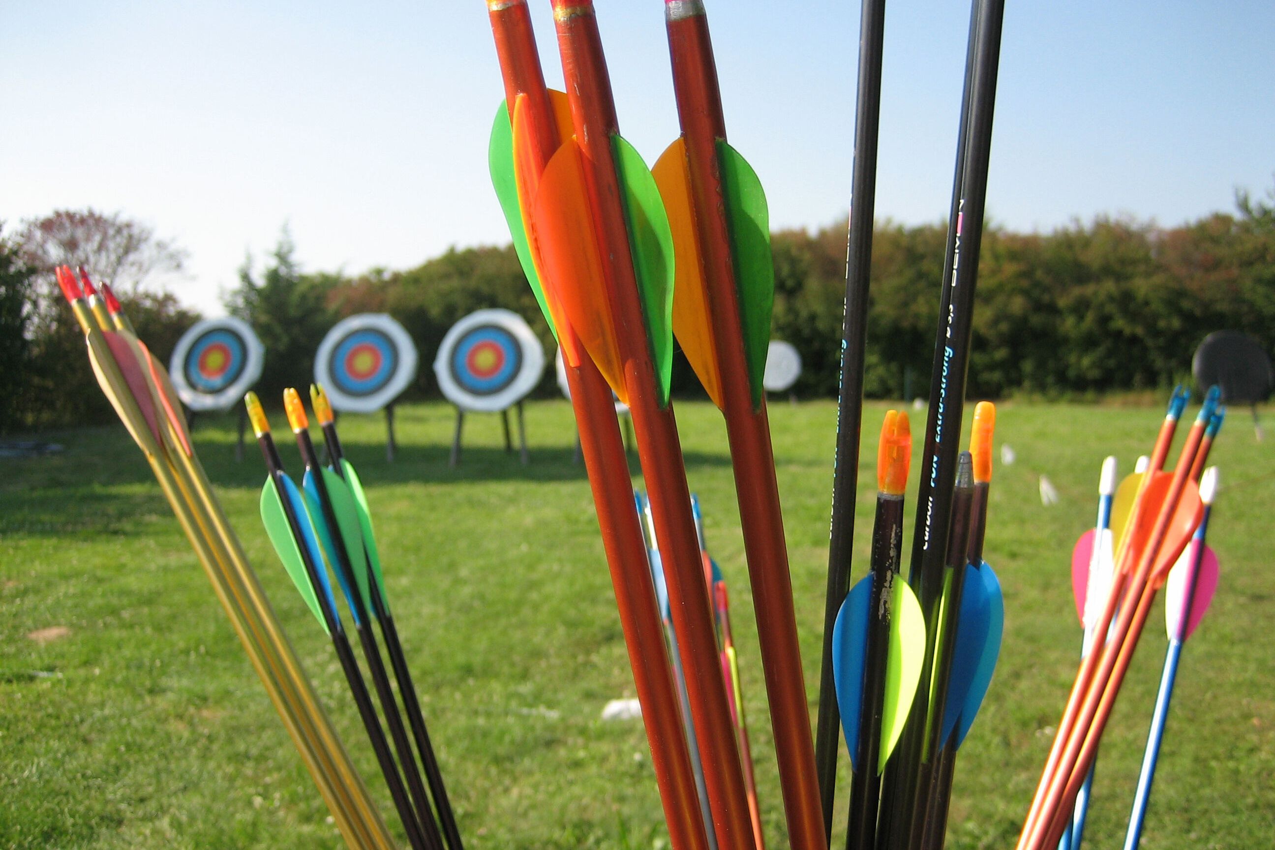 Das Bild zeigt eine Reihe von Pfeilen, die in einem Halter stecken. Die Pfeile haben verschiedene Farben, darunter Orange, Grün, Gelb, Blau, Schwarz und Rot. Im Hintergrund sind mehrere Zielscheiben in einem Rasenbereich zu sehen. Der Himmel ist blau.