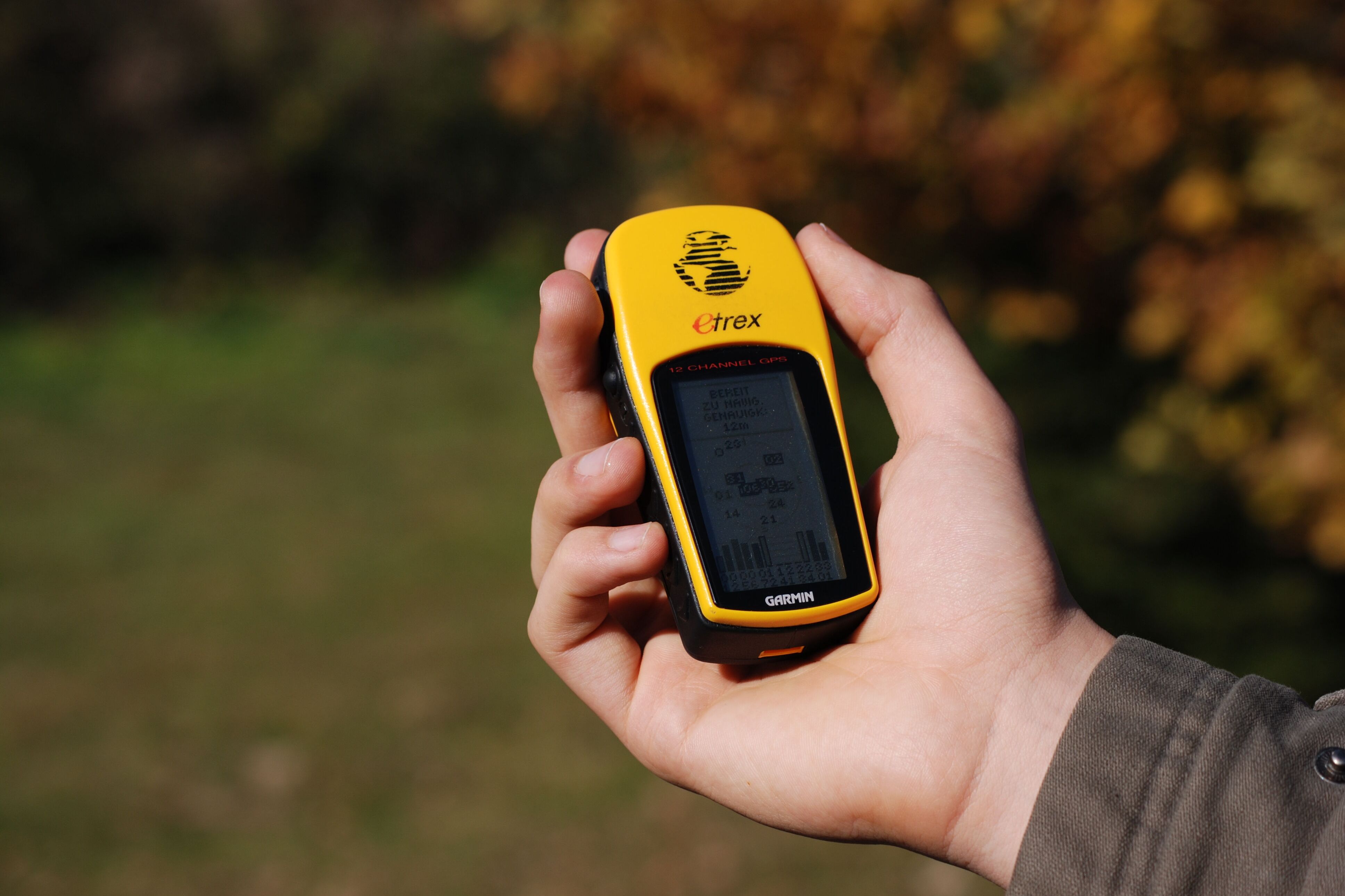 Ein Mann hält ein gelbes GPS-Gerät in seiner Hand. Der Bildschirm des Geräts ist schwarz und zeigt Informationen an. Im Hintergrund ist ein verschwommener Blick auf Bäume mit braunen und grünen Blättern zu sehen. Der Mann trägt ein braunes Hemd.