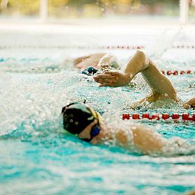 Das Bild zeigt eine Schwimmszene. Im Vordergrund sind zwei Schwimmer in einer Schwimmspur zu sehen, die sich im Kraulstil im Wasser bewegen. Die Schwimmer tragen Schwimmbrillen und Badekappen. Einer der Schwimmer hat einen Arm über dem Wasser und den anderen im Wasser. Im Hintergrund sind weitere Schwimmer zu sehen. Das Wasser ist blau und es gibt rote Bojen, die die Schwimmbahnen markieren. Der Hintergrund ist unscharf und zeigt eine helle Decke.