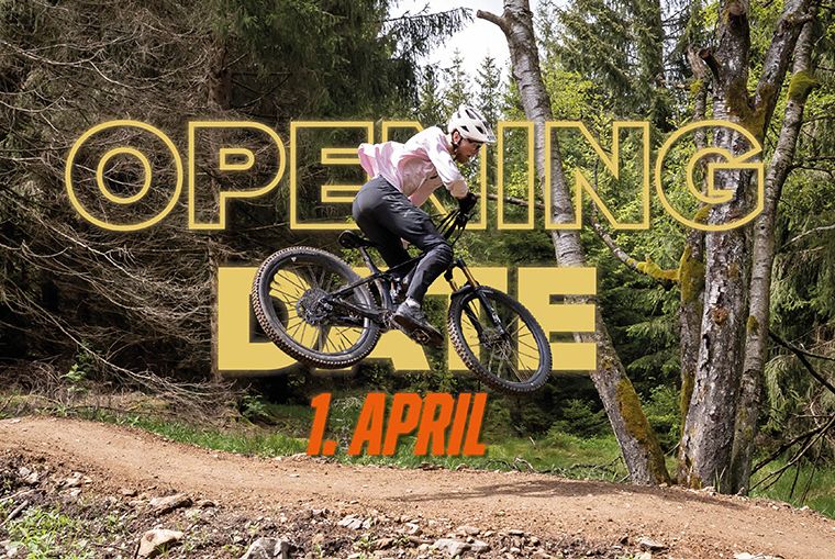 Mountainbiker springt über eine Schanze mit Text "OPENING DATE 1. APRIL" im Wald.