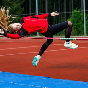 Das Bild zeigt ein Mädchen, das Hochsprung macht. Es ist in der Luft und versucht, über die Latte zu springen. Sie trägt eine rote Jacke, schwarze Leggings und Laufschuhe. Ihre Haare sind blond und fliegen in der Luft. Die Latte ist rot und grün. Das Bild wird auf einer roten Sportbahn vor einem grünen Hintergrund aufgenommen. Unter der Latte befindet sich eine blaue Matte.