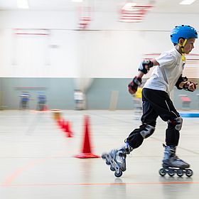Das Bild zeigt einen Jungen, der auf Inlineskates in einer Halle fährt. Er ist mit einem blauen Helm, Knieschützern, Ellbogenschützern und Handschuhen geschützt. Er trägt ein weißes T-Shirt und eine schwarze Hose. Er fährt mit hoher Geschwindigkeit, was durch die Bewegungsunschärfe im Hintergrund angezeigt wird. Auf dem Boden sind rote Kegel aufgereiht, die wahrscheinlich einen Parcours bilden.