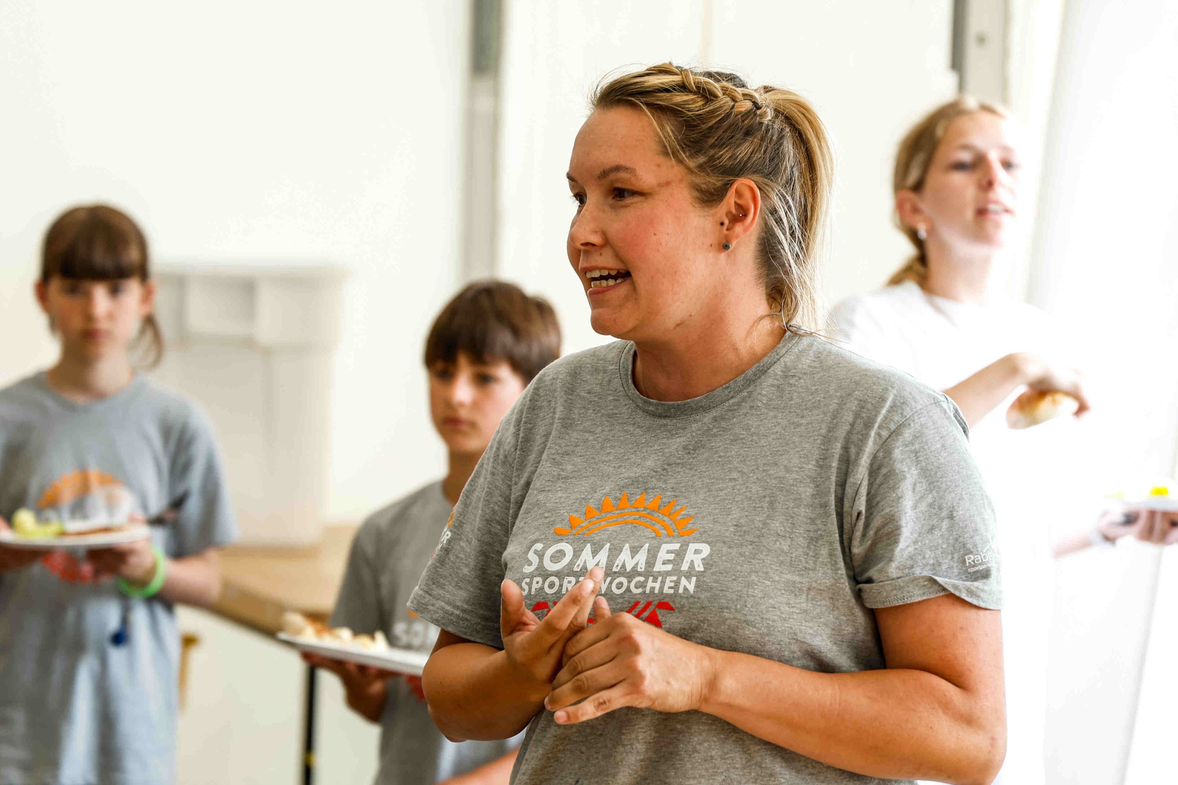 Das Foto zeigt eine Gruppe von Menschen, die in einem hellen Raum stehen. Im Vordergrund steht eine Frau mit blonden Haaren, die zu einem Zopf geflochten sind. Sie trägt ein graues T-Shirt mit dem Aufdruck "SOMMER SPORTWOCHEN". Sie scheint zu sprechen und ihre Hände sind in einer Geste vor ihrer Brust verschränkt. Hinter ihr stehen mehrere weitere Personen, die teilweise unscharf dargestellt sind. Zwei Kinder, ein Junge und ein Mädchen, stehen links und halten Teller mit Essen. Eine weitere Frau steht rechts im Hintergrund.