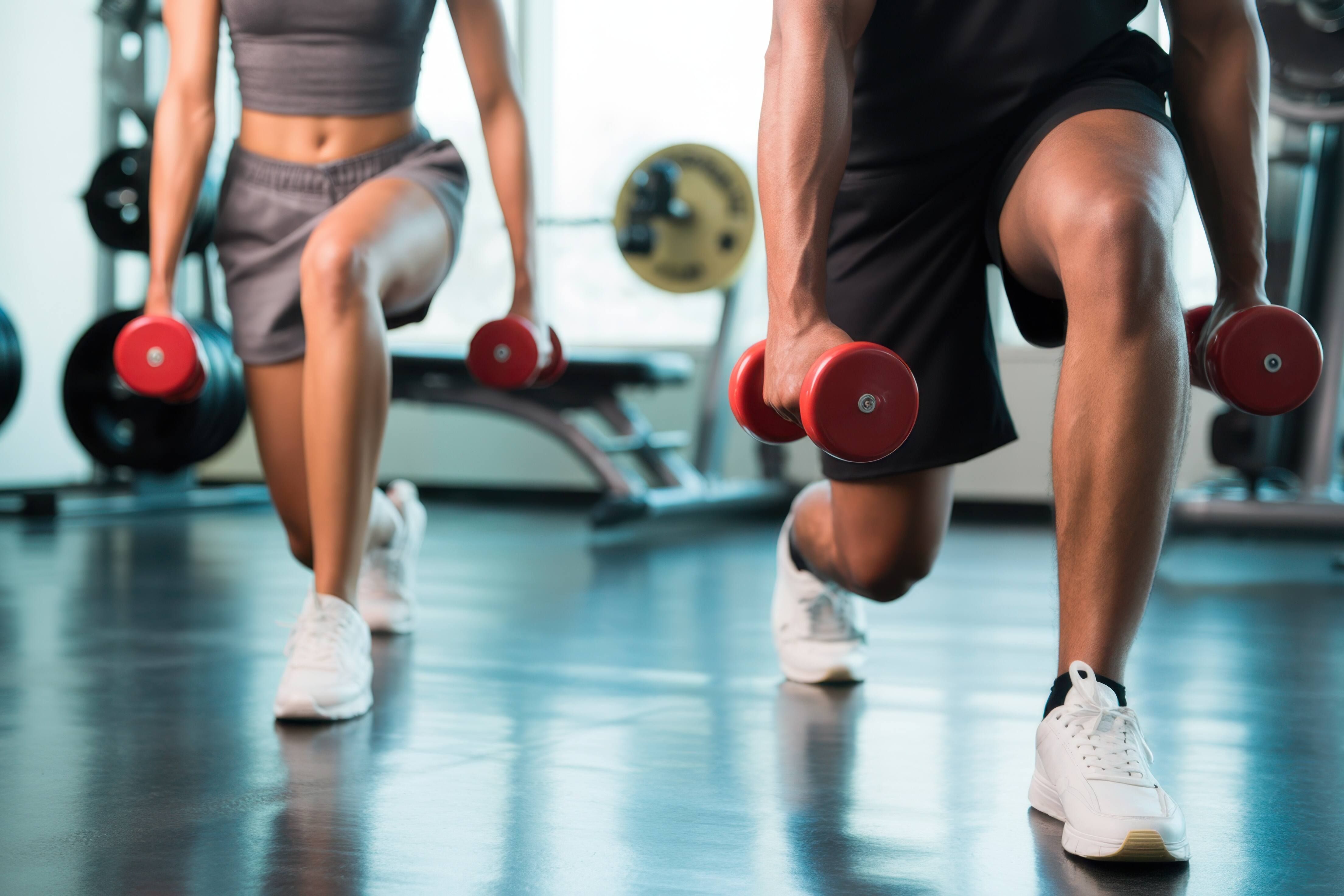 Das Bild zeigt zwei Personen, die in einem Fitnessstudio Ausfallschritte machen. Beide Personen halten in jeder Hand eine rote Kurzhantel. Die Person im Vordergrund ist ein Mann, dessen Beine und Arme zu sehen sind, er trägt weiße Turnschuhe und schwarze Shorts. Die andere Person im Hintergrund ist eine Frau, deren Oberkörper und Beine zu sehen sind, sie trägt graue Shorts und ein graues Top und weiße Turnschuhe. Im Hintergrund sind Fitnessgeräte zu sehen.