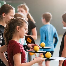 Das Bild zeigt eine Gruppe von Kindern in einer Sporthalle. Sie scheinen gerade eine Sportart zu spielen, bei der sie kleine gelbe Bälle mit Schlägern schlagen. Im Vordergrund stehen mehrere Mädchen die jeweils einen Ball auf dem Schläger balancieren. Eines der Mädchen in einem roten T-Shirt hält einen Schläger in der Hand und legt gerade einen gelben Ball auf den Schläger. Andere Mädchen halten ebenfalls Schläger und Bälle. Im Hintergrund sind weitere Kinder zu sehen, die in verschiedene Richtungen schauen oder sich bewegen.