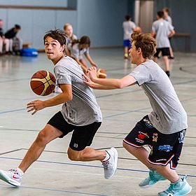 Das Bild zeigt zwei Jungen, die in einer Turnhalle Basketball spielen. Der Junge im Vordergrund dribbelt den Ball mit der rechten Hand und blickt nach links. Er trägt ein graues T-Shirt, schwarze Shorts und weiße Turnschuhe mit roten Akzenten. Der Ball ist orange-braun mit schwarzen Rillen. Der andere Junge versucht, ihm den Ball abzunehmen und streckt seine Hand nach dem ersten Jungen aus. Er trägt ein graues T-Shirt, Basketballshorts mit Teamlogos und hellblaue Turnschuhe. Im Hintergrund sind weitere Personen zu sehen, einige davon scheinen ebenfalls Basketball zu spielen, während andere zuschauen. Sie tragen ähnliche Outfits wie die beiden Jungen im Vordergrund. Auf dem Boden sind Linien für das Spielfeld aufgezeichnet.