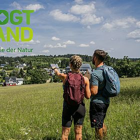 Sportpark Rabenberg EURORANDO 2026 Aussteller: Tourismusverband Vogtland; © Dirk Rückschloß | pixore photography Ein Paar mit Rucksäcken blickt auf ein Dorf und Bergpanorama. Linke obere Ecke Logo "Vogtland Sinfonie der Natur"