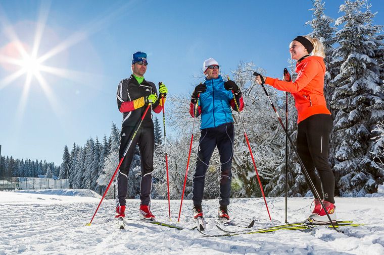 Das Foto zeigt drei Personen auf Langlaufskiern in einer verschneiten Winterlandschaft. Im Hintergrund leuchten Bäume, der Himmel ist blau. Die Sonne scheint hell und wirft Sonnenstrahlen. Die drei Skifahrer sind in Sportkleidung gekleidet und scheinen sich zu unterhalten oder eine Pause zu machen.