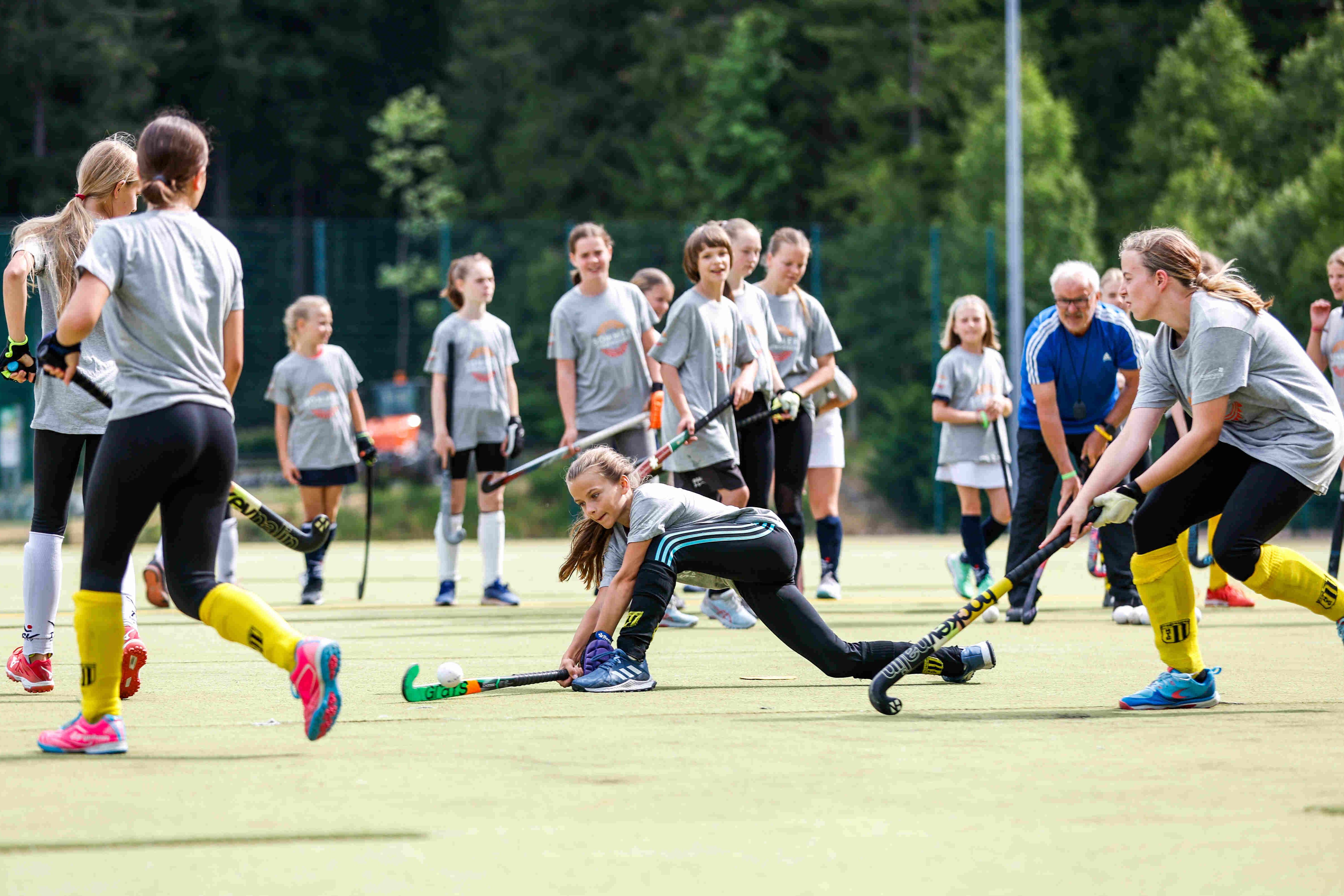 Das Bild zeigt eine Gruppe junger Mädchen, die Hockey spielen. Sie tragen alle graue T-Shirts, schwarze Leggings und farbige Socken und Schuhe. Einige Mädchen tragen gelbe Stutzen. Einige Mädchen haben ihre Haare zu einem Pferdeschwanz zusammengebunden. Auf dem grünen Rasen stehen auch einige Mädchen und beobachten das Spiel. Im Hintergrund stehen Bäume und ein älterer Mann in einem blauen T-Shirt. Das Wetter ist sonnig. Ein Mädchen ist in einer niedrigen Position abgebildet, während es versucht, den Hockeyball mit seinem Schläger zu treffen. Die anderen Mädchen stehen im Hintergrund und beobachten das Spiel. Der Mann im blauen T-Shirt scheint ein Trainer zu sein. 