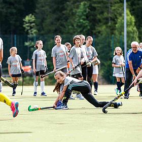 Das Bild zeigt eine Gruppe junger Mädchen, die Hockey spielen. Sie tragen alle graue T-Shirts, schwarze Leggings und farbige Socken und Schuhe. Einige Mädchen tragen gelbe Stutzen. Einige Mädchen haben ihre Haare zu einem Pferdeschwanz zusammengebunden. Auf dem grünen Rasen stehen auch einige Mädchen und beobachten das Spiel. Im Hintergrund stehen Bäume und ein älterer Mann in einem blauen T-Shirt. Das Wetter ist sonnig. Ein Mädchen ist in einer niedrigen Position abgebildet, während es versucht, den Hockeyball mit seinem Schläger zu treffen. Die anderen Mädchen stehen im Hintergrund und beobachten das Spiel. Der Mann im blauen T-Shirt scheint ein Trainer zu sein. 