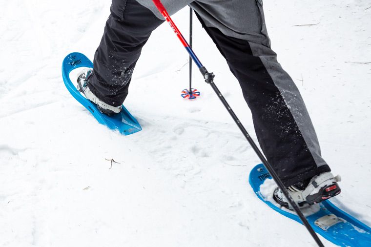 Das Bild zeigt eine Person, die Schneeschuhe in tiefem Schnee trägt. Die Person trägt dunkle Hosen und weiße Schuhe. Die Schneeschuhe sind blau. In der Szene sind auch Skistöcke vorhanden.