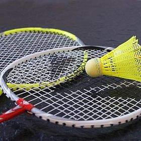 Das Bild zeigt zwei Badmintonschläger und einen gelben Federball. Die Schläger sind in einer diagonalen Anordnung angeordnet, wobei sich der vordere Schläger über dem hinteren befindet. Der Griff des vorderen Schlägers ist rot und weiß, während der Griff des hinteren Schlägers gelb ist. Die Rahmen der Schläger sind silberfarben. Das Netz der Schläger ist weiß. Der Federball liegt auf dem Netz des vorderen Schlägers. Der Federball hat einen gelben Federkopf und einen hellgelben Korkfuß. Der Hintergrund des Bildes ist dunkel.