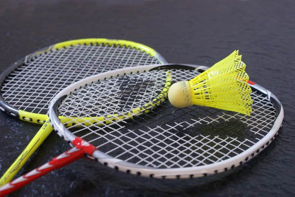 Das Bild zeigt zwei Badmintonschläger und einen gelben Federball. Die Schläger sind in einer diagonalen Anordnung angeordnet, wobei sich der vordere Schläger über dem hinteren befindet. Der Griff des vorderen Schlägers ist rot und weiß, während der Griff des hinteren Schlägers gelb ist. Die Rahmen der Schläger sind silberfarben. Das Netz der Schläger ist weiß. Der Federball liegt auf dem Netz des vorderen Schlägers. Der Federball hat einen gelben Federkopf und einen hellgelben Korkfuß. Der Hintergrund des Bildes ist dunkel.