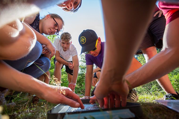 Das Bild zeigt eine Gruppe von Menschen, die sich um etwas auf dem Boden versammelt haben. Aus der Perspektive des Betrachters blickt man von unten nach oben in die Szene hinein. Im Zentrum des Geschehens liegt vermutlich eine flache Schale oder ein Tablett, an dem mehrere Personen arbeiten. Die Personen sind jung und tragen Freizeitkleidung. Einige haben die Köpfe gesenkt, um sich die Arbeit anzusehen. Eine Person trägt eine Sonnenbrille und eine Baseballkappe. Im Hintergrund ist etwas Grün zu sehen, vermutlich Gras oder Bäume, und der Himmel ist blau.