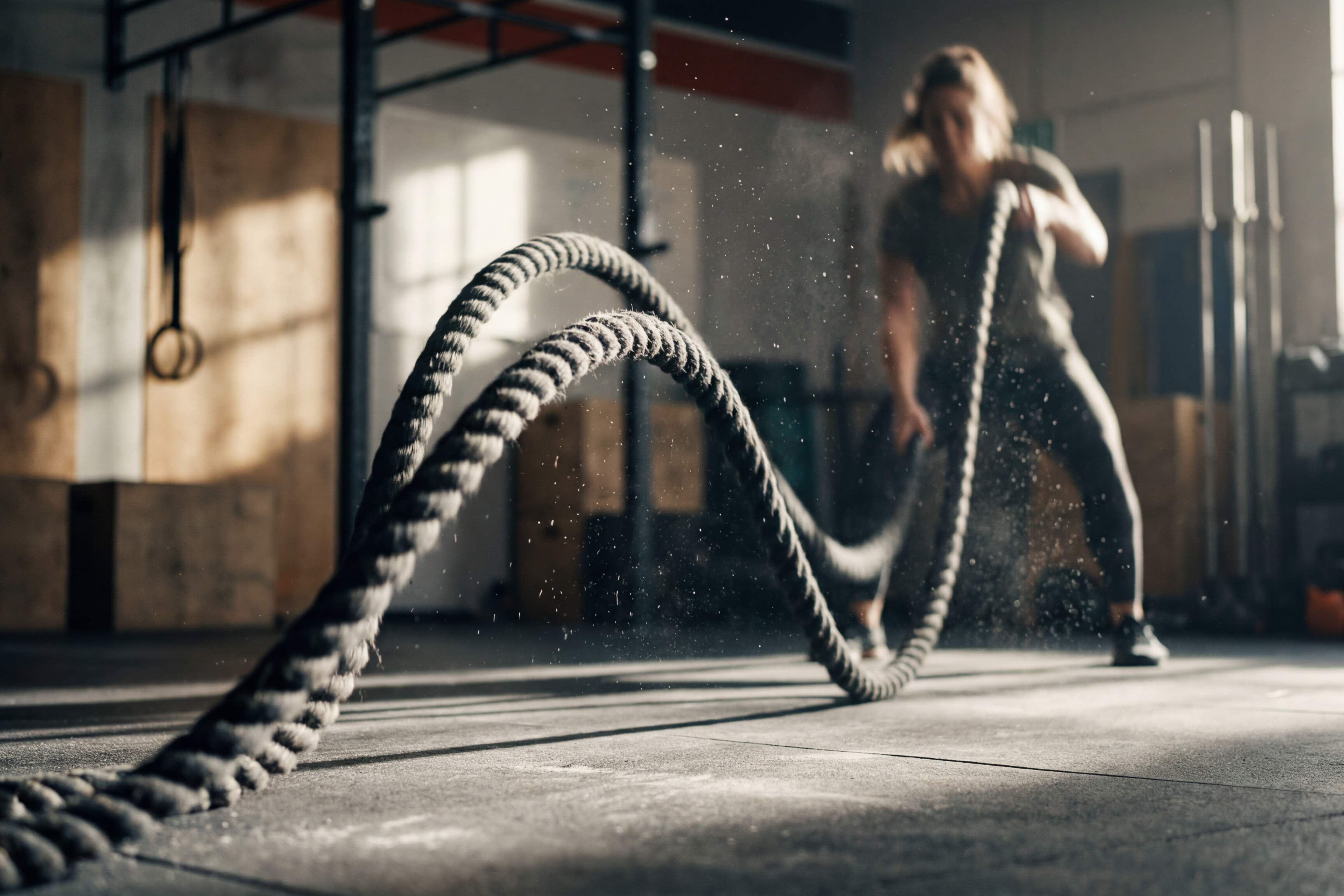 Das Bild zeigt eine unscharfe Person in einem Fitnessstudio, die trainiert. Sie hält zwei Seile in den Händen, eins in jeder Hand und scheint diese im Wechsel schnell auf und ab zu bewegen, sodass Staubpartikel durch die Luft wirbeln. Der Hintergrund ist unscharf, aber es ist möglich, Geräte wie Gewichte und ein Fenster zu sehen.