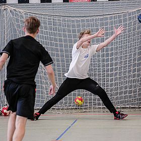 Das Bild zeigt eine Szene aus einem Handballspiel. Im Vordergrund sind zwei Jungen zu sehen. Einer der Jungen steht mit dem Rücken zur Kamera und trägt ein schwarzes T-Shirt und eine schwarze Hose. Er scheint gerade einen Wurf auszuführen. Der andere Junge steht im Tor und versucht, den Ball zu fangen. Er trägt ein weißes T-Shirt und eine schwarze Hose. Er hat die Arme weit geöffnet und versucht, den Ball abzuwehren. Der Ball ist gerade im Flug und nähert sich dem Tor. Im Hintergrund ist ein Handballtor zu sehen, das aus einem Netz und einem Rahmen besteht. Auf dem Boden liegen weitere Bälle. Die Halle hat graue Wände.