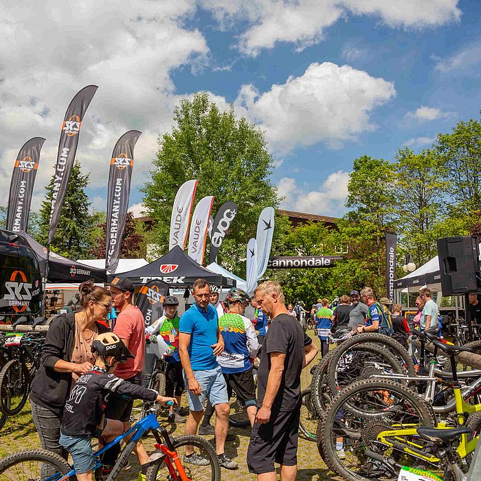 Das Foto zeigt eine Outdoor-Veranstaltung, vermutlich ein Fahrradrennen oder eine Messe. Im Vordergrund sind viele Menschen zu sehen, die sich um Fahrräder herum versammeln. Es gibt eine große Auswahl an Mountainbikes, die an Ständern ausgestellt sind. Einige Leute scheinen die Fahrräder zu begutachten oder sich mit den Anbietern zu unterhalten. Im Hintergrund sind mehrere Zelte und Werbebanner zu sehen. Einige der Banner sind hoch und wehen im Wind. Der Himmel ist blau und mit einigen Wolken bedeckt, was auf einen sonnigen Tag hindeutet. Bäume und Büsche umgeben den Veranstaltungsort, was eine natürliche Umgebung schafft. Insgesamt vermittelt das Bild den Eindruck einer lebendigen und gut besuchten Veranstaltung im Freien, bei der sich alles um Fahrräder dreht.
