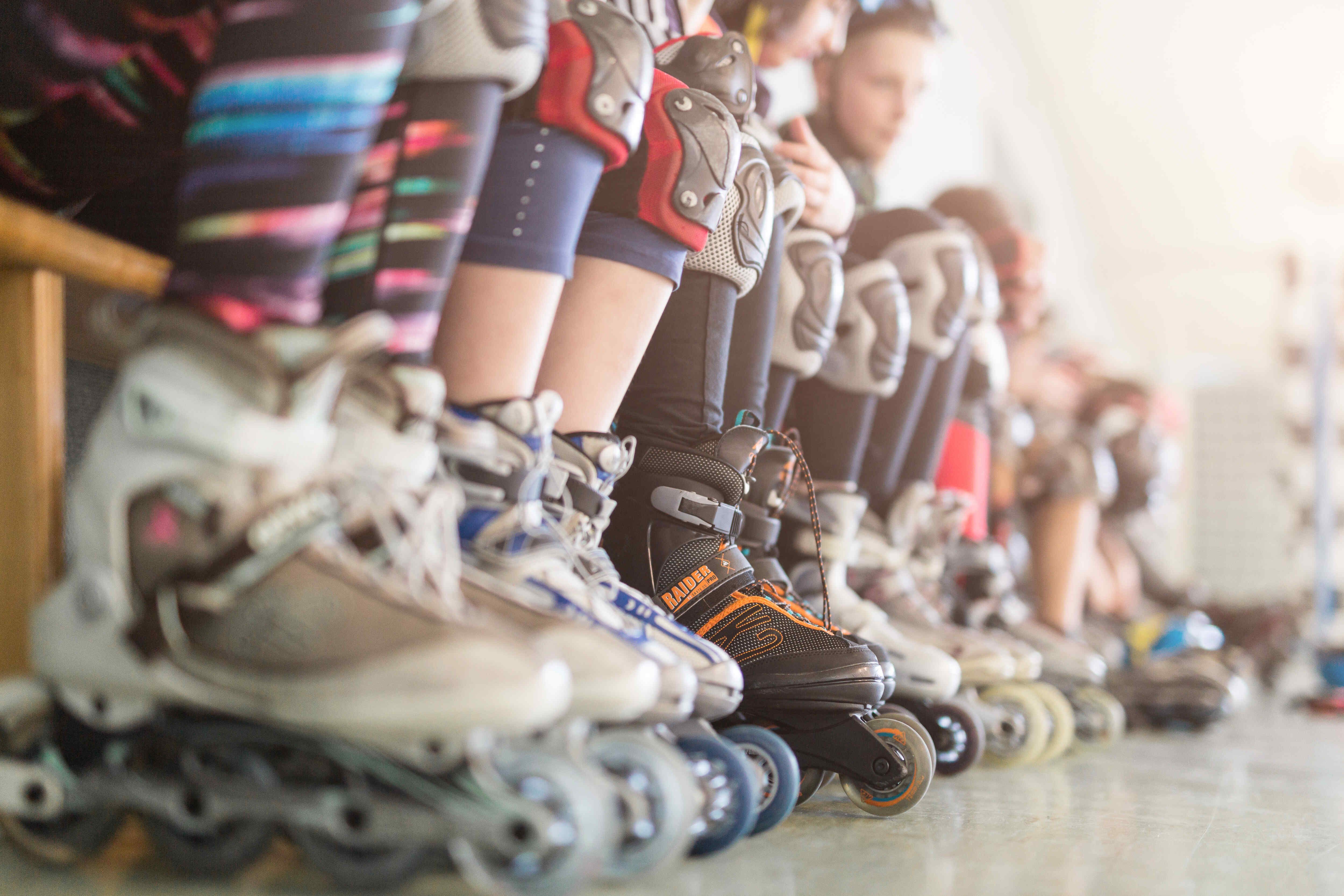 Das Bild zeigt eine Reihe von Inline-Skates in einer Reihe. Die Skates sind in verschiedenen Farben gehalten und stehen auf einer hellen Oberfläche. Die Füße der Skater sind teilweise zu sehen, wobei Schutzkleidung wie Knieschützer und Strumpfhosen getragen werden. Der Hintergrund ist unscharf, was den Fokus auf die Inline-Skates lenkt.