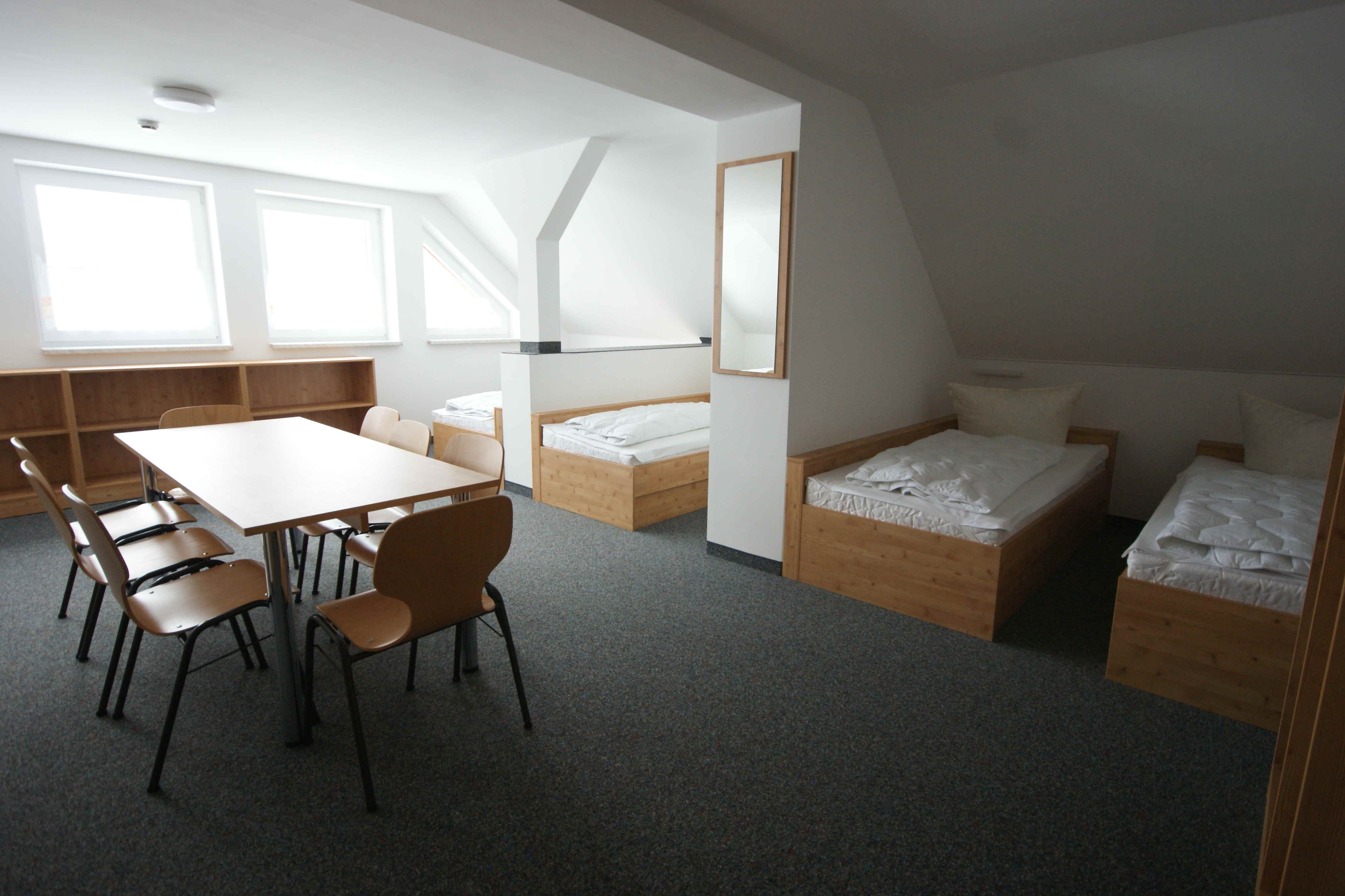 Das Bild zeigt einen Innenraum, der wie ein Schlafsaal oder ein Gästezimmer aussieht. Der Raum ist hell und mit neutralen Farben gehalten. Im Vordergrund befindet sich ein Tisch mit sechs Stühlen. Der Tisch ist rechteckig und hat eine helle, glatte Oberfläche. Die Stühle sind aus Holz und haben schwarze Metallbeine. Im Hintergrund befinden sich mehrere Betten. Die Betten sind aus Holz und haben weiße Bettwäsche. Es gibt auch einen Spiegel an der Wand. Der Boden ist mit einem grauen Teppich ausgelegt. Die Wände sind weiß.