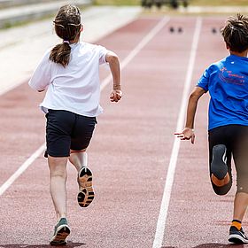 Das Bild zeigt zwei Kinder, die auf einer roten Laufbahn rennen. Beide sind von hinten zu sehen. Das Mädchen, das links auf dem Bild rennt, trägt ein weißes T-Shirt und schwarze Shorts. Sie hat ihr Haar zu einem Pferdeschwanz zusammengebunden. Ihre Arme sind angewinkelt. Der Junge, der rechts auf dem Bild rennt, trägt ein blaues T-Shirt mit einem Logo auf der Rückseite. Er trägt schwarze Shorts und schwarze Schuhe mit orangefarbenen Socken. Seine Arme sind angewinkelt. Die Laufbahn hat weiße Markierungen.