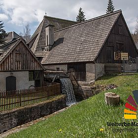 Sportpark_Rabenberg EURORANDO 2026 Aussteller: Welterbe Montanregion Erzgebirge; Historisches Gebäude mit Wasserrad, Wasserfall und Schindeldach, umgeben von Bäumen und Grünflächen.