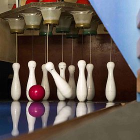 Das Bild zeigt eine Kegelbahn. Im Vordergrund stehen Kegel, die zum Teil durch die rote Kegelkugel umgeworfen werden. Die Kegel sind weiß und haben die typische Form. Die Kegelkugel ist rot und liegt auf der Bahn. Die Kegelbahn ist blau und spiegelt die Kegel und die Kugel wider.