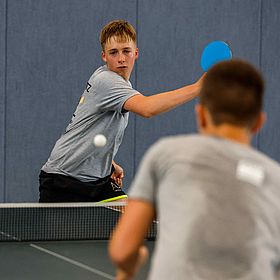 Das Foto zeigt zwei Jungen, die Tischtennis spielen. Der Junge im Vordergrund ist unscharf und von hinten zu sehen. Er trägt ein graues T-Shirt. Der andere Junge, der im Fokus steht, ist auch mit einem grauen T-Shirt bekleidet und schaut in Richtung der Kamera. Er hat einen blonden Haarschnitt. Er hält einen blauen Schläger in der Hand und schlägt gerade den Ball. Der Ball ist auch im Fokus und befindet sich in der Mitte des Bildes. Im Hintergrund befindet sich eine blaue Wand und ein Tischtennisnetz.