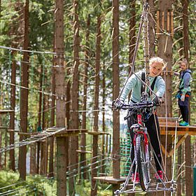 Das Bild zeigt eine junge Frau, die mit einem Fahrrad über einen Hochseilgarten fährt. Sie ist in einer grünen Jacke und schwarzen Hosen gekleidet und trägt Handschuhe. Sie hat ein breites Lächeln im Gesicht und schaut in die Kamera. Das Fahrrad ist rot und hat dicke Reifen. Im Hintergrund ist ein anderer junger Mensch zu sehen, der auf einer Plattform steht und ebenfalls einen Gurt trägt. Beide sind von Bäumen umgeben, und es gibt eine Reihe von Hindernissen, die durch den Hochseilgarten führen.