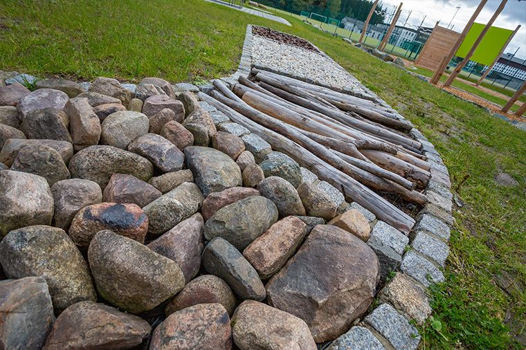 Das Bild zeigt einen Barfußpfad im Freien. Im Vordergrund liegt ein Feld aus großen, runden Natursteinen, die dicht nebeneinander angeordnet sind und eine unebene Trittfläche bilden. Daneben befindet sich ein weiteres Feld mit schmalen, parallel verlegten Holzleisten, die eine gleichmäßige, gerillte Oberfläche haben. Der Barfußpfad ist in grünen Rasen eingebettet, im Hintergrund sind Bäume und weitere Pfadabschnitte erkennbar.