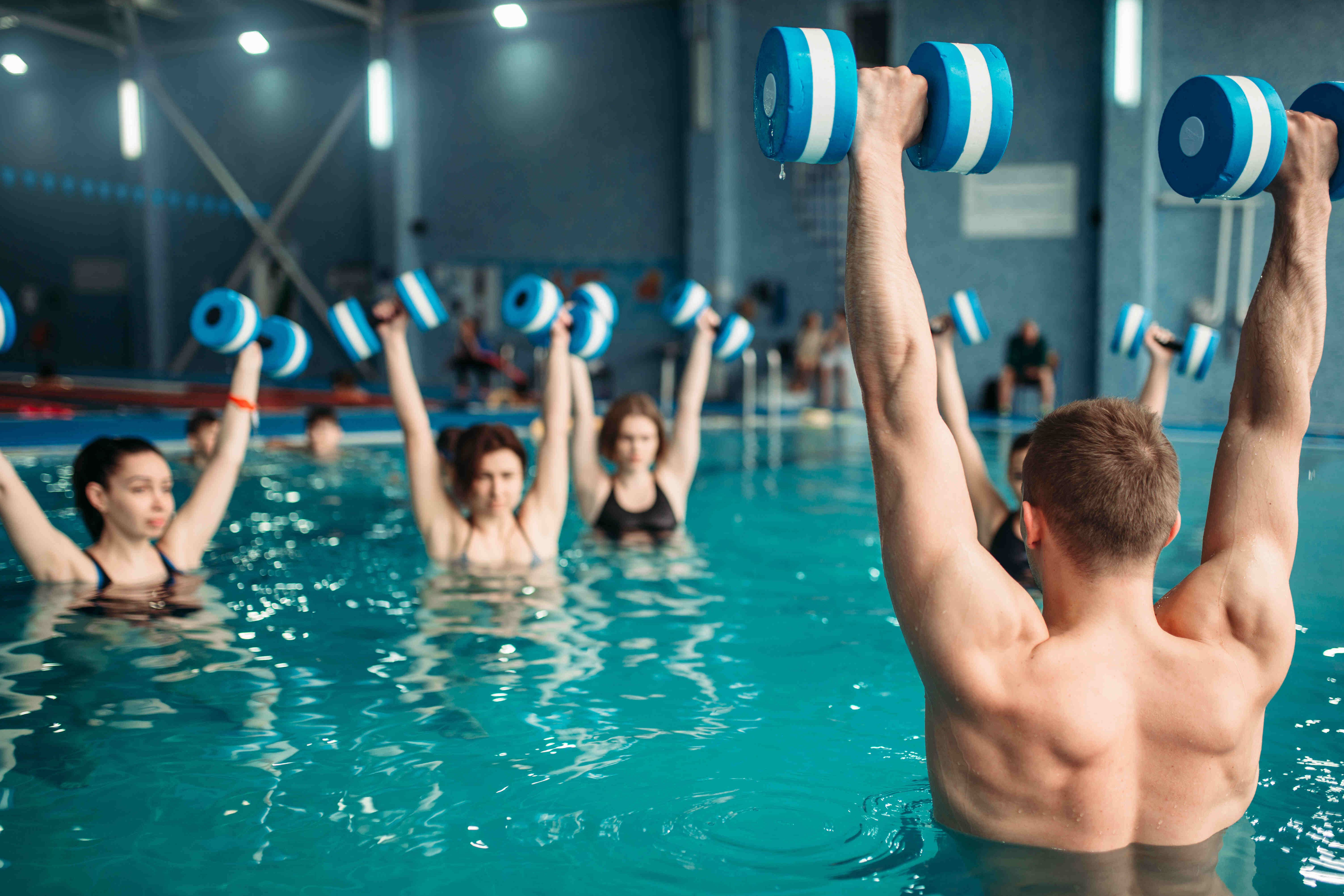Das Bild zeigt eine Gruppe von Menschen, die im Wasser trainieren. Sie halten blaue Hanteln hoch in die Luft. Die Personen sind im Wasser bis zur Brust. Der Hintergrund ist eine Schwimmhalle mit blauen Wänden und einigen Zuschauern.