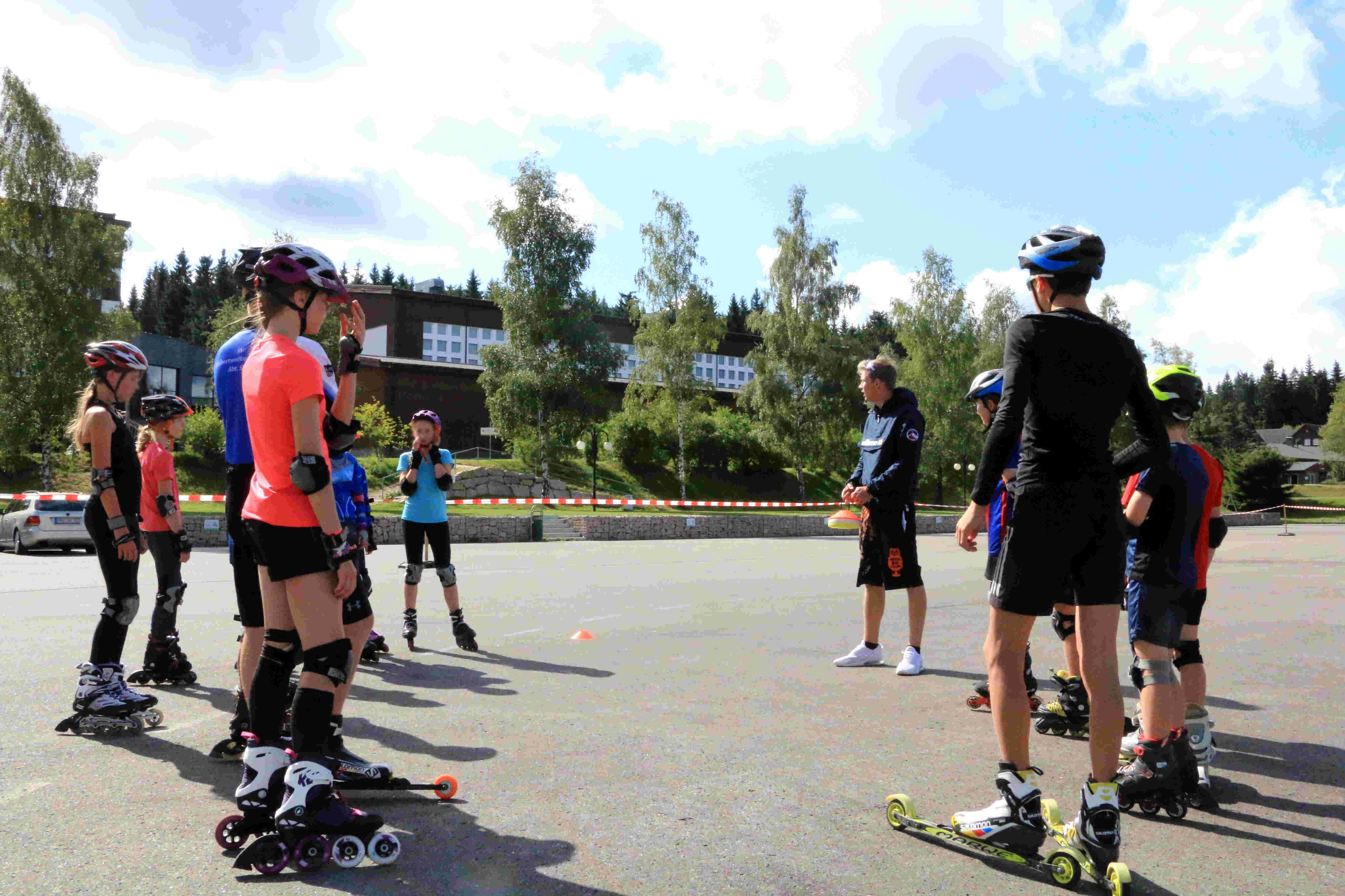 Das Bild zeigt eine Gruppe von Menschen, die auf einer asphaltierten Fläche Inlineskaten oder Rollski fahren. Die Gruppe besteht aus Kindern und Jugendlichen, sowie einem Mann. Die meisten tragen Helme, Knieschützer und Ellbogenschützer, was darauf hindeutet, dass sie sich auf Sicherheit konzentrieren. Im Hintergrund sind Bäume und Gebäude zu sehen, was darauf hindeutet, dass die Szene im Freien stattfindet. Der Himmel ist blau und sonnig. Die Leute scheinen sich in einem Training oder einer Unterrichtsstunde zu befinden, da ein Mann in der Mitte der Gruppe steht und Anweisungen zu geben scheint.