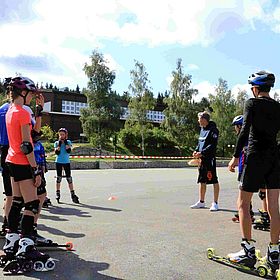 Das Bild zeigt eine Gruppe von Menschen, die auf einer asphaltierten Fläche Inlineskaten oder Rollski fahren. Die Gruppe besteht aus Kindern und Jugendlichen, sowie einem Mann. Die meisten tragen Helme, Knieschützer und Ellbogenschützer, was darauf hindeutet, dass sie sich auf Sicherheit konzentrieren. Im Hintergrund sind Bäume und Gebäude zu sehen, was darauf hindeutet, dass die Szene im Freien stattfindet. Der Himmel ist blau und sonnig. Die Leute scheinen sich in einem Training oder einer Unterrichtsstunde zu befinden, da ein Mann in der Mitte der Gruppe steht und Anweisungen zu geben scheint.