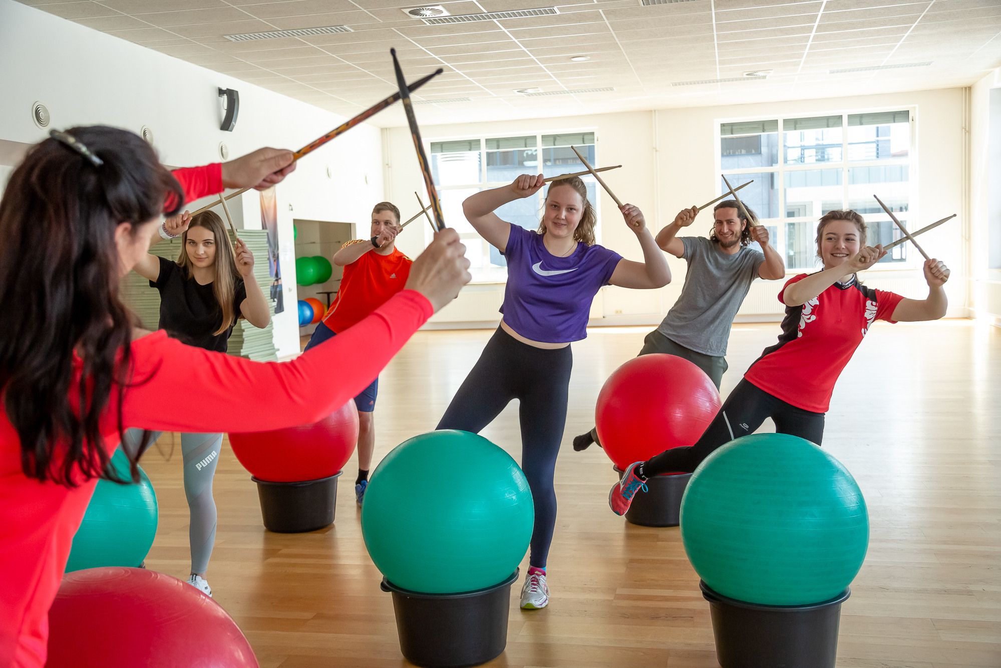 Das Bild zeigt eine Gruppe von sechs Personen in einem Fitnessstudio. Alle halten Drumsticks in den Händen und stehen anscheinend über Gymnastikbällen. Die Personen sind in Sportkleidung gekleidet und scheinen in Bewegung zu sein. Im Hintergrund sind Fenster und weitere Gymnastikbälle zu sehen. Die Atmosphäre wirkt lebendig und sportlich.