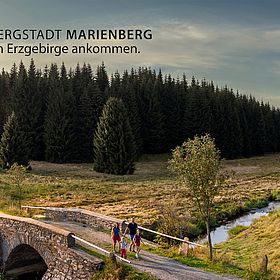 Sportpark Rabenberg EURORANDO 2026 Aussteller: Stadt Marienberg; © Uwe Meinhold Familie überquert Steinbrücke im Erzgebirge neben einem Bach und einem Wald; Logo Stadt Marienberg in oberer linker Ecke