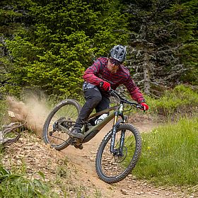 Sportpark Rabenberg: TrailCenter®-Rabenberg Das Bild zeigt einen Mountainbiker, der einen Bergpfad hinunterfährt. Der Radfahrer trägt einen Helm, eine Sonnenbrille, ein rot-weißes Langarmshirt, schwarze Hosen und rote Handschuhe. Er ist auf einem Mountainbike, das sich in Bewegung befindet. Der Hintergrund des Bildes zeigt eine grüne Waldumgebung. Der Pfad, auf dem der Radfahrer fährt, ist aus brauner Erde mit einigen Steinen und Wurzeln. Der Radfahrer befindet sich in der Mitte des Bildes und bewegt sich nach unten und links. Das Hinterrad des Bikes wirbelt etwas Staub auf. Die Gesamtstimmung des Bildes ist sportlich und actionreich.