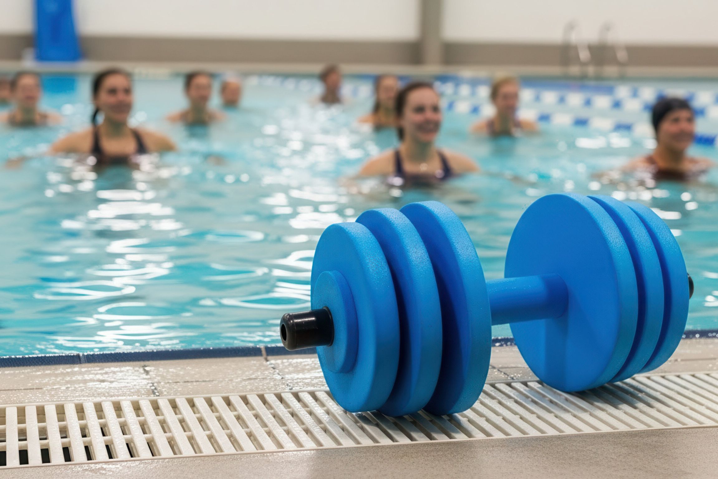 Das Bild zeigt eine Gruppe von Menschen, die Wassergymnastik machen. Im Vordergrund befindet sich eine blaue Hantel. Im Hintergrund befinden sich die Personen im Wasser.