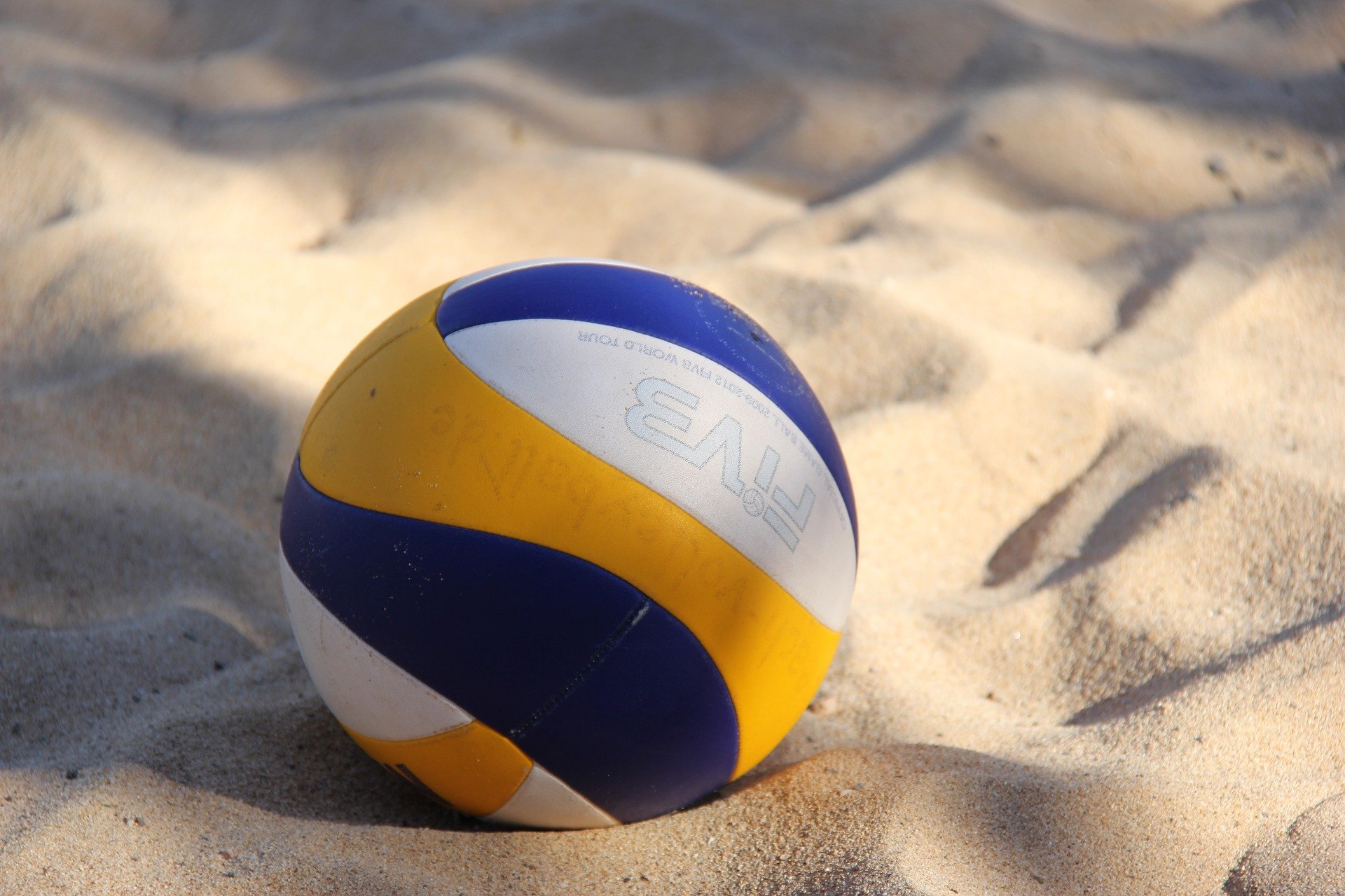 Das Bild zeigt einen Volleyball, der im Sand liegt. Der Ball ist eine Kombination aus Blau, Weiß und Gelb. Das Logo "FIVB" ist auf dem Ball zu sehen. Der Sand ist hell und weist eine unebene Textur auf.