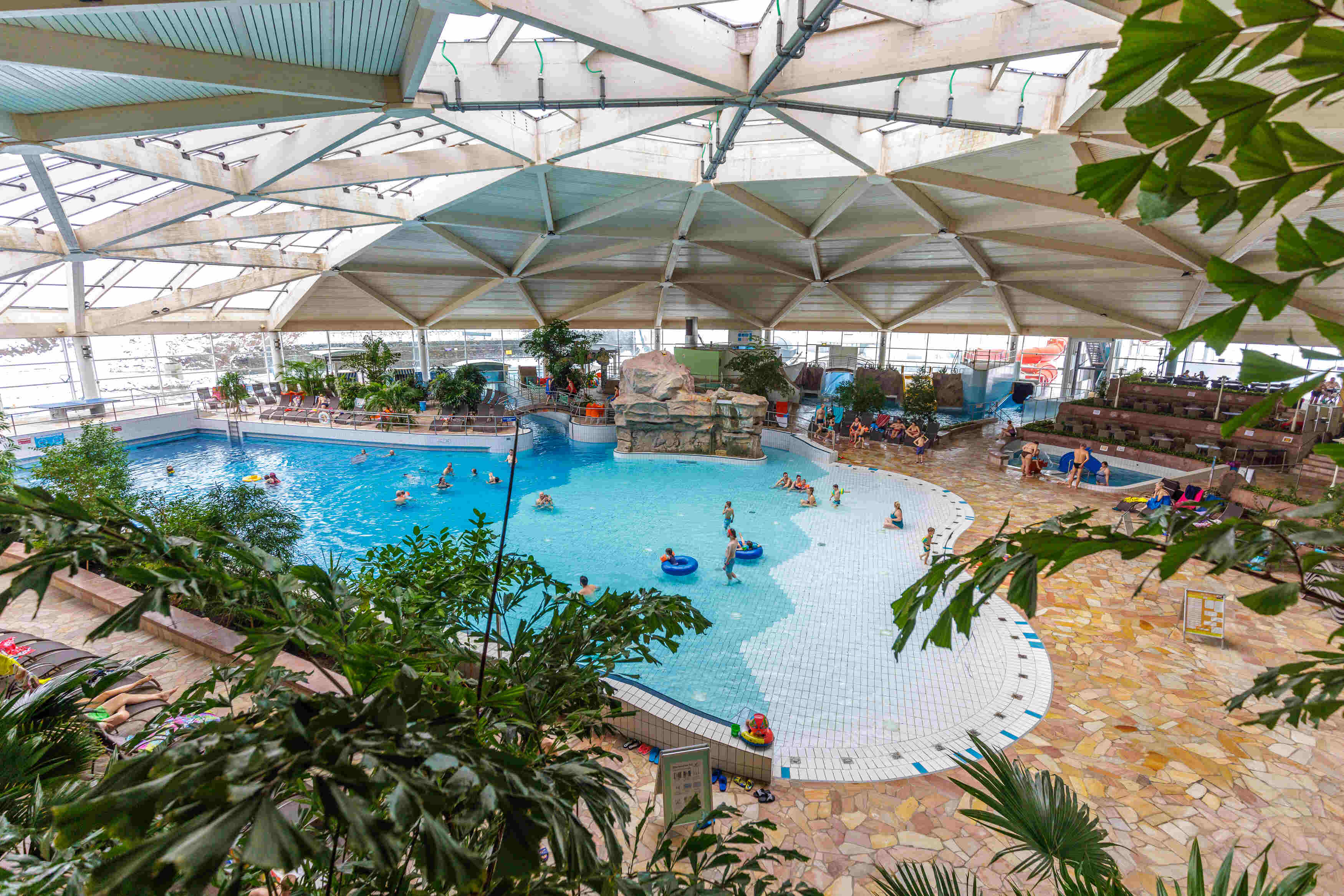 Das Bild zeigt eine Indoor-Badelandschaft. Im Vordergrund sind grüne Blätter zu sehen, die einen Teil der Szene verbergen. Im Mittelpunkt des Bildes befindet sich ein großes Schwimmbecken mit blauem Wasser. Es gibt mehrere Personen im Becken, die schwimmen oder sich entspannen. Einige von ihnen benutzen Schwimmreifen. Das Becken hat eine unregelmäßige Form und ist von einem weißen Boden mit blauen Fliesen umgeben. Um das Schwimmbecken herum befinden sich verschiedene Bereiche. Auf der rechten Seite gibt es einen Bereich mit Liegestühlen, auf denen sich einige Leute entspannen. Es gibt auch einen Bereich mit einem Felsen und einer Brücke, der wie ein Wasserfall aussieht. Weiter rechts ist eine Rutsche zu sehen. Die Decke des Schwimmbades ist aus Glas und Holz gebaut und hat eine interessante geometrische Form. Durch die Decke fällt Tageslicht in den Raum.