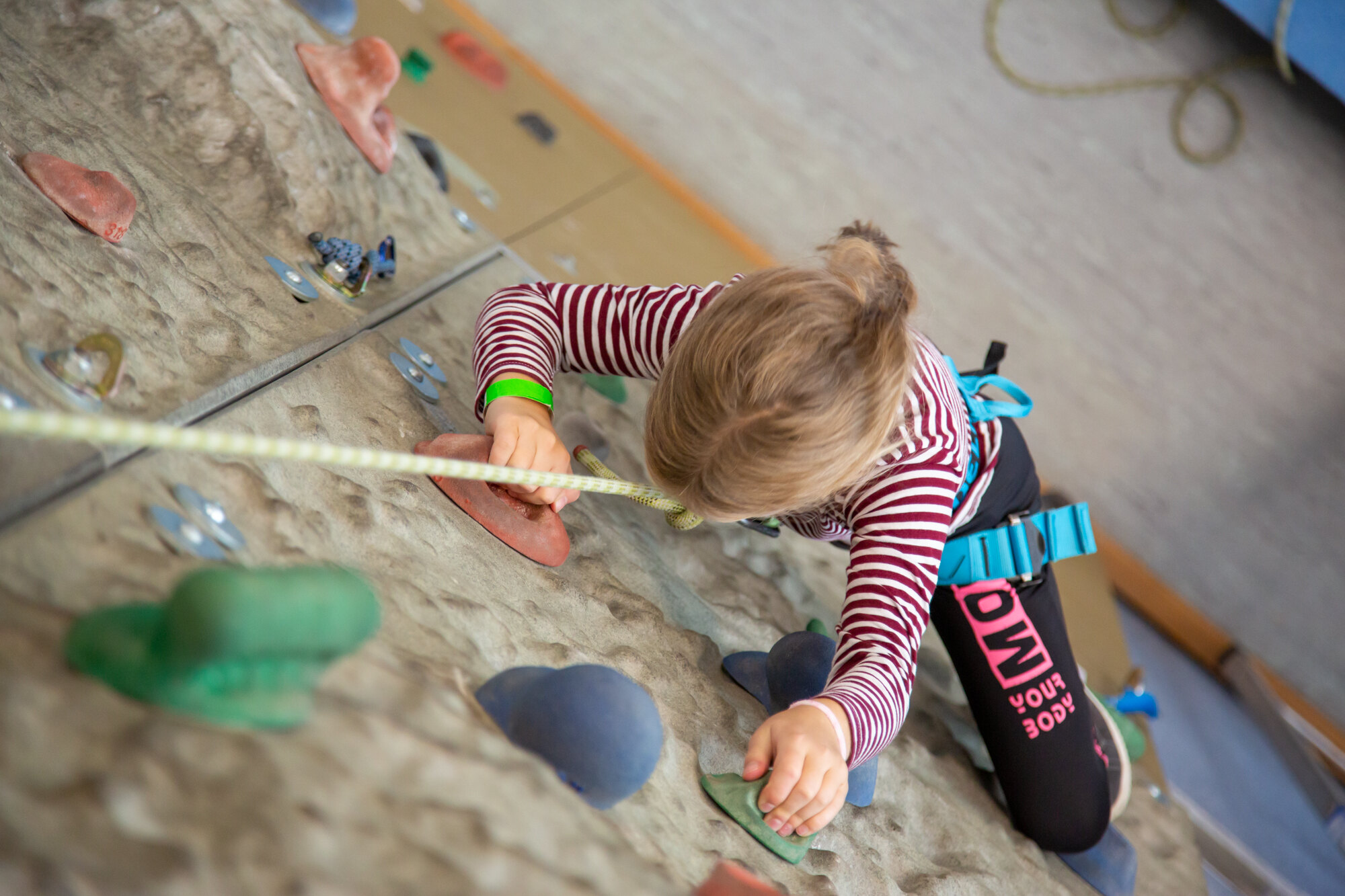 Das Bild zeigt ein Kind beim Klettern an einer Kletterwand. Das Kind ist von oben fotografiert, so dass man es von Kopf bis Fuß sehen kann, während es die Wand erklimmt. Es ist ein junges Mädchen, dessen blondes Haar zu einem kleinen Pferdeschwanz zusammengebunden ist. Sie trägt ein langärmliges rot-weiß gestreiftes Oberteil, schwarze Leggings und einen blauen Klettergurt. Ein Sicherungsseil ist an dem Klettergurt des Kindes befestigt und verläuft nach oben und aus dem Bild heraus.