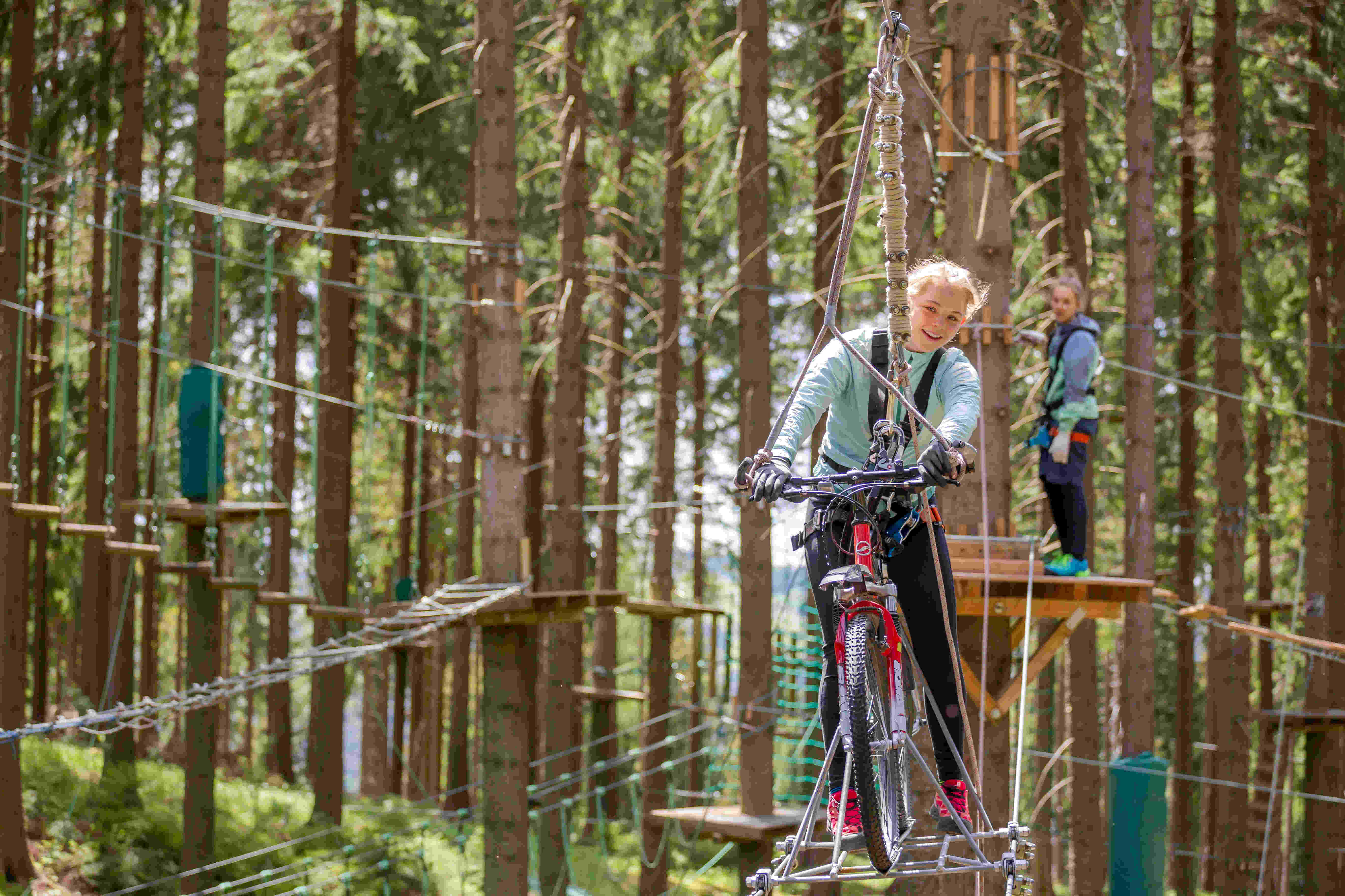 Das Bild zeigt eine junge Frau, die mit einem Fahrrad über einen Hochseilgarten fährt. Sie ist in einer grünen Jacke und schwarzen Hosen gekleidet und trägt Handschuhe. Sie hat ein breites Lächeln im Gesicht und schaut in die Kamera. Das Fahrrad ist rot und hat dicke Reifen. Im Hintergrund ist ein anderer junger Mensch zu sehen, der auf einer Plattform steht und ebenfalls einen Gurt trägt. Beide sind von Bäumen umgeben, und es gibt eine Reihe von Hindernissen, die durch den Hochseilgarten führen.