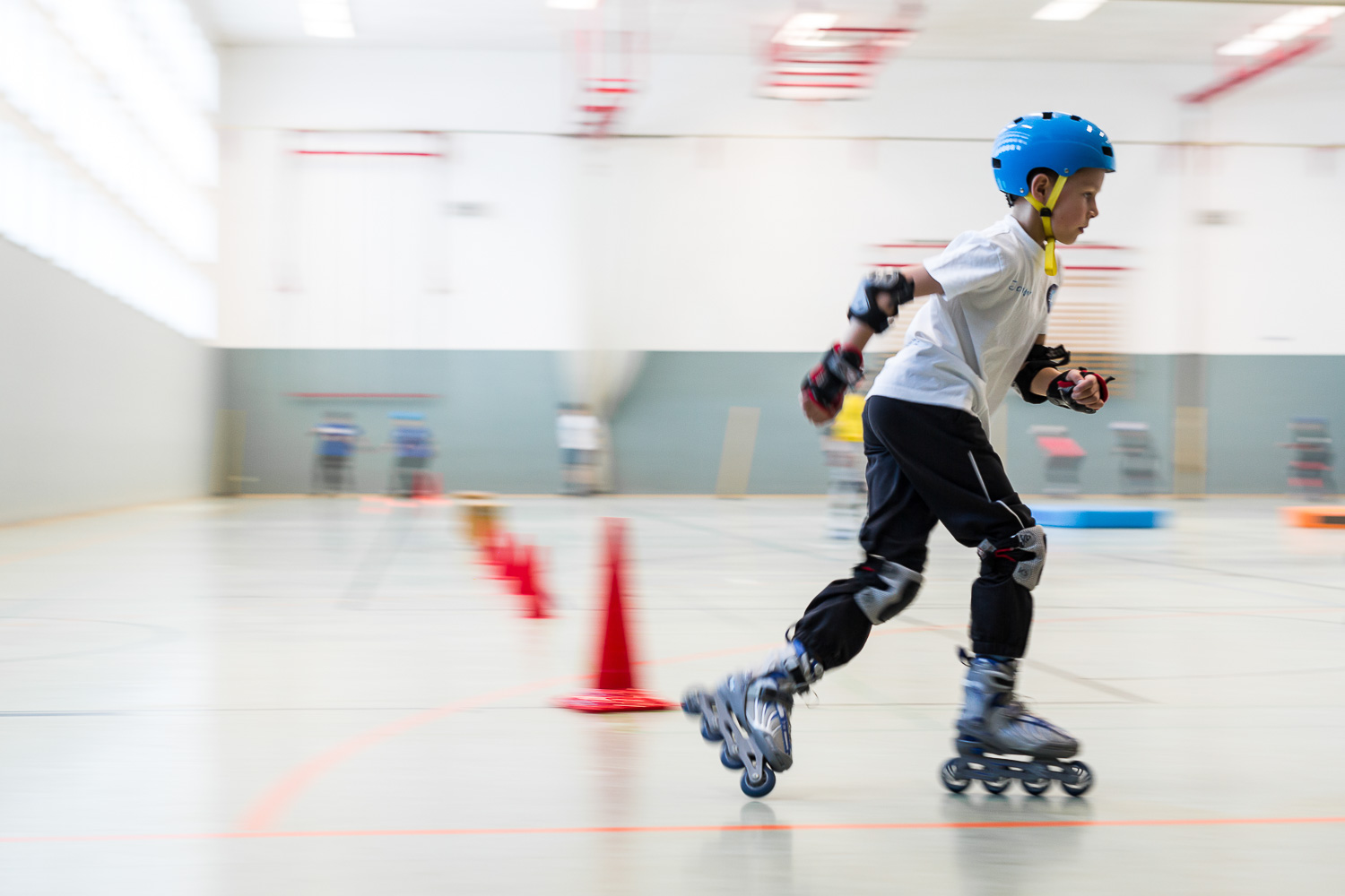 Das Bild zeigt einen Jungen, der auf Inlineskates in einer Halle fährt. Er ist mit einem blauen Helm, Knieschützern, Ellbogenschützern und Handschuhen geschützt. Er trägt ein weißes T-Shirt und eine schwarze Hose. Er fährt mit hoher Geschwindigkeit, was durch die Bewegungsunschärfe im Hintergrund angezeigt wird. Auf dem Boden sind rote Kegel aufgereiht, die wahrscheinlich einen Parcours bilden.
