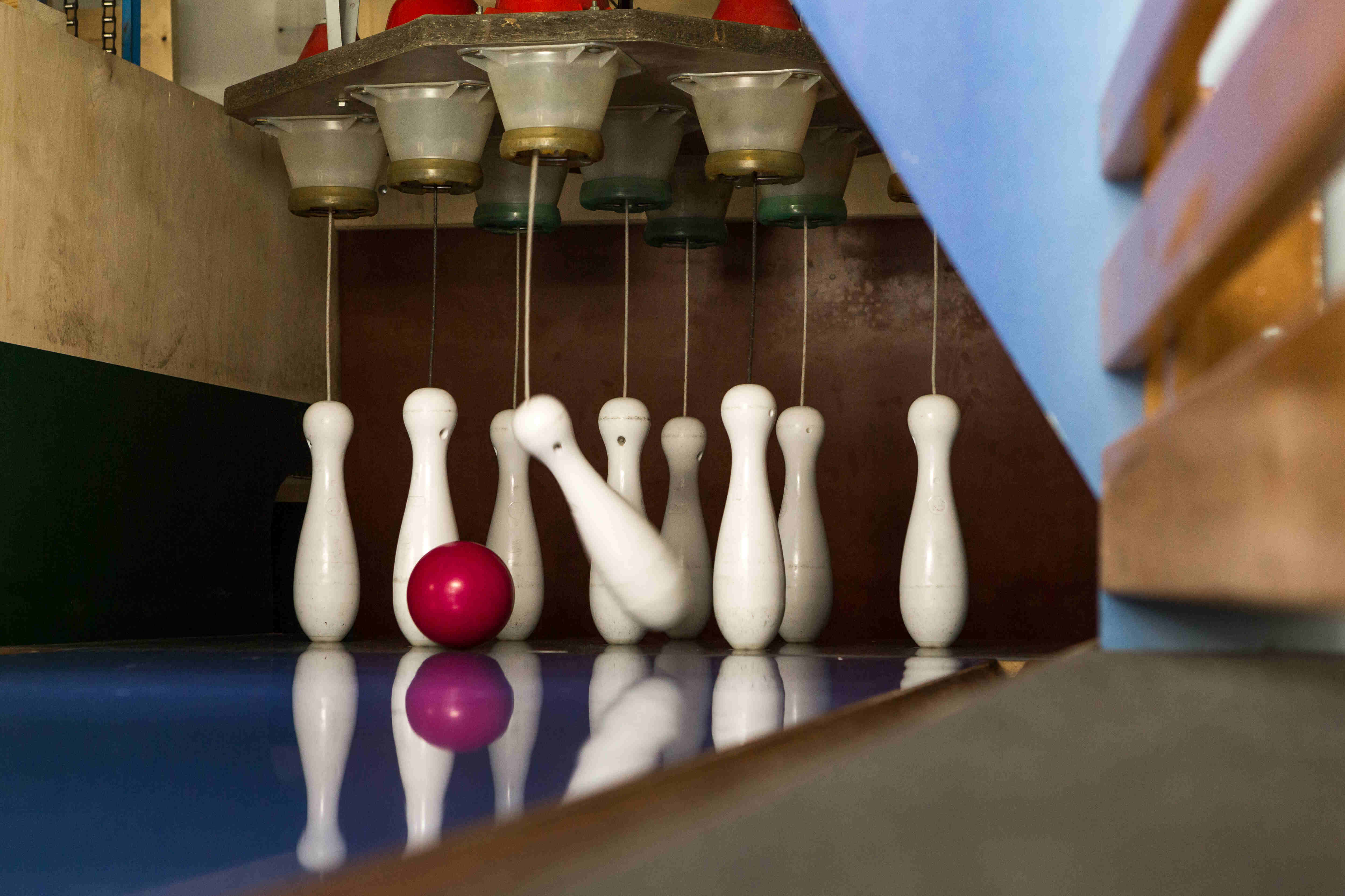 Das Bild zeigt eine Kegelbahn. Im Vordergrund stehen Kegel, die zum Teil durch die rote Kegelkugel umgeworfen werden. Die Kegel sind weiß und haben die typische Form. Die Kegelkugel ist rot und liegt auf der Bahn. Die Kegelbahn ist blau und spiegelt die Kegel und die Kugel wider.