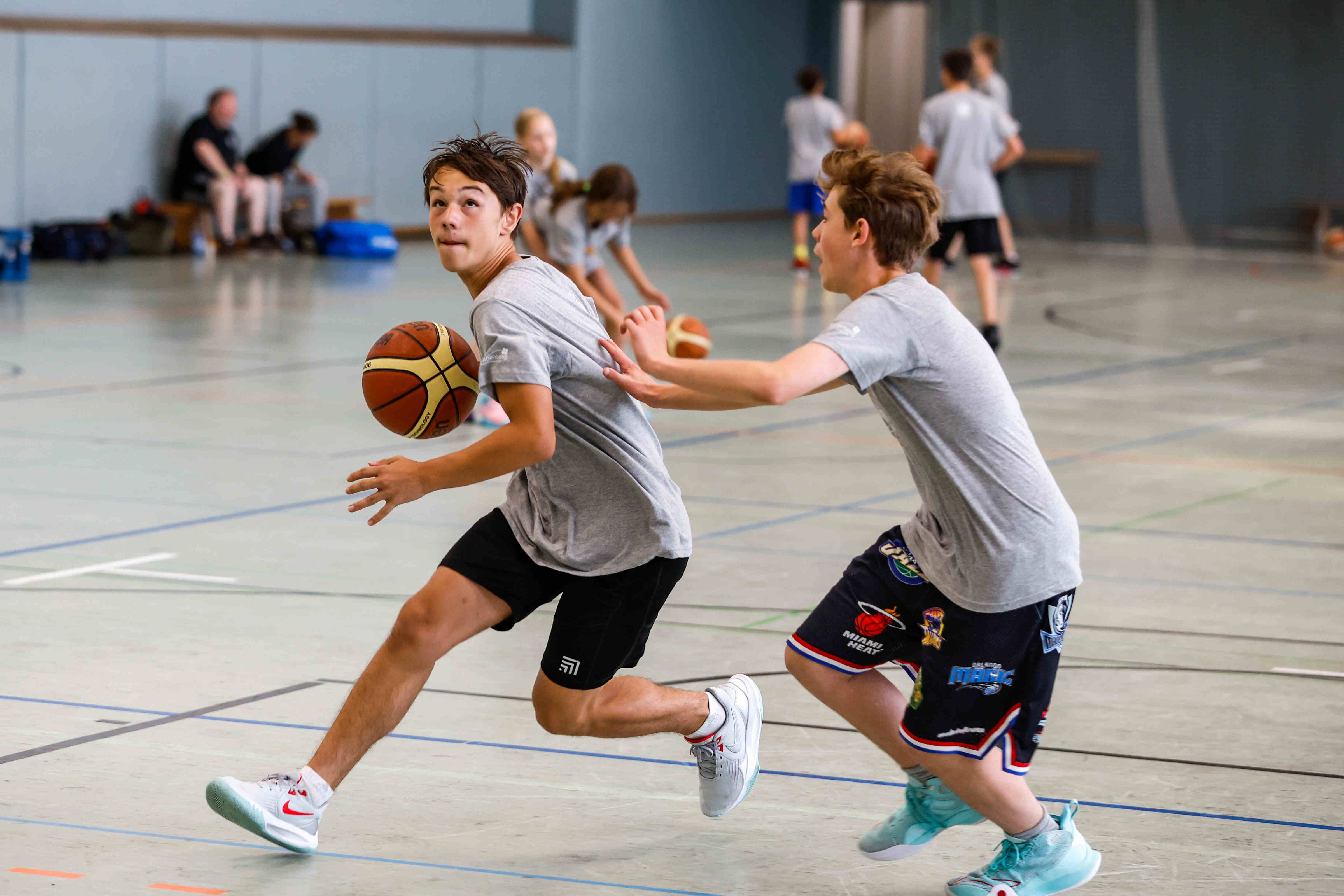 Das Bild zeigt zwei Jungen, die in einer Turnhalle Basketball spielen. Der Junge im Vordergrund dribbelt den Ball mit der rechten Hand und blickt nach links. Er trägt ein graues T-Shirt, schwarze Shorts und weiße Turnschuhe mit roten Akzenten. Der Ball ist orange-braun mit schwarzen Rillen. Der andere Junge versucht, ihm den Ball abzunehmen und streckt seine Hand nach dem ersten Jungen aus. Er trägt ein graues T-Shirt, Basketballshorts mit Teamlogos und hellblaue Turnschuhe. Im Hintergrund sind weitere Personen zu sehen, einige davon scheinen ebenfalls Basketball zu spielen, während andere zuschauen. Sie tragen ähnliche Outfits wie die beiden Jungen im Vordergrund. Auf dem Boden sind Linien für das Spielfeld aufgezeichnet.