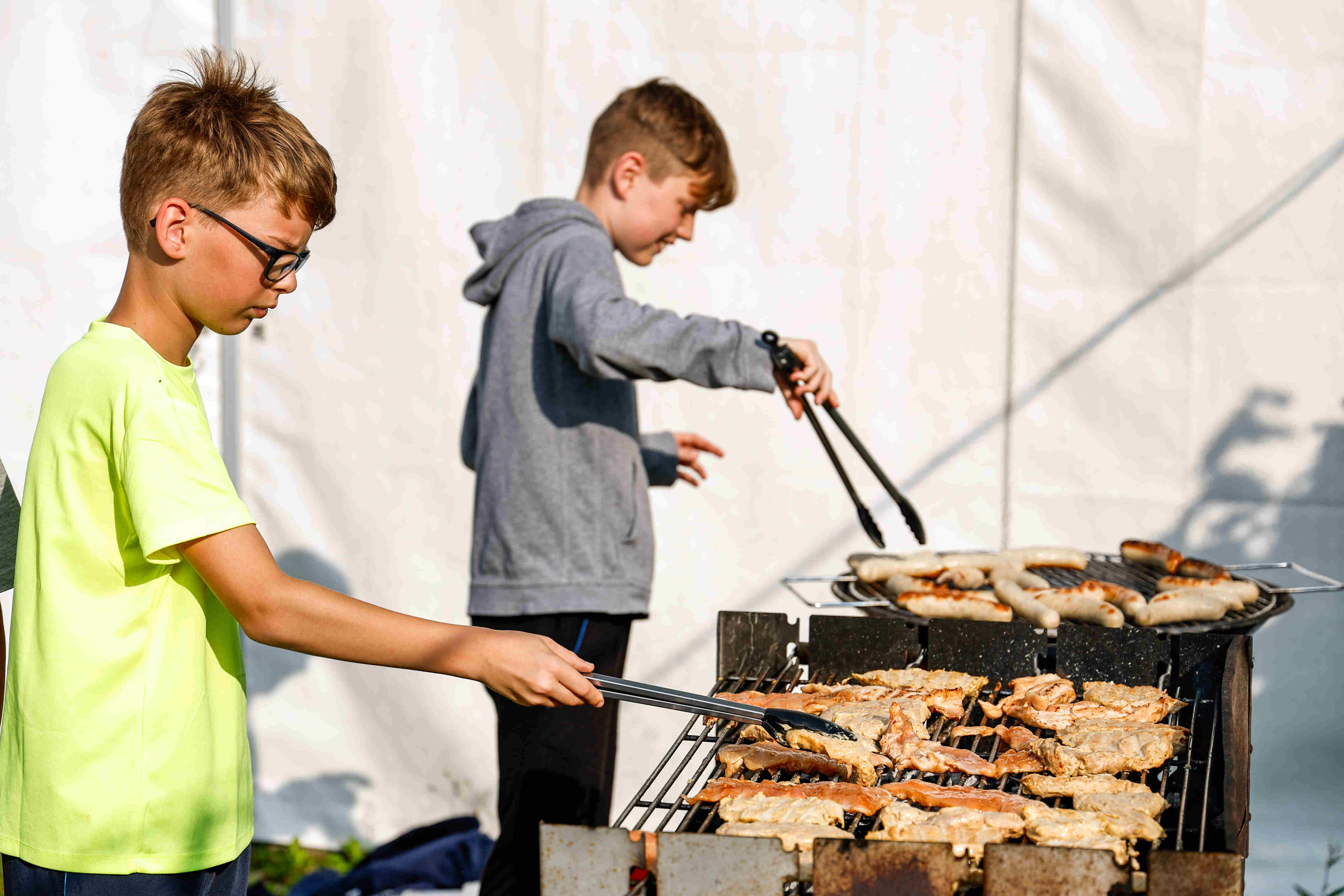 Das Bild zeigt zwei Jungen, die im Freien grillen. Der Junge auf der linken Seite trägt ein neongelbes T-Shirt und eine Brille. Er hält eine Zange und scheint Fleisch auf dem Grill zu wenden. Der andere Junge, der sich im Hintergrund befindet, trägt einen grauen Kapuzenpullover und scheint ebenfalls mit der Grillzange zu hantieren. Auf dem Grill liegen Würste und Fleischstücke. Der Hintergrund ist ein weißes Zelt. Die Szene wirkt sommerlich und entspannt.